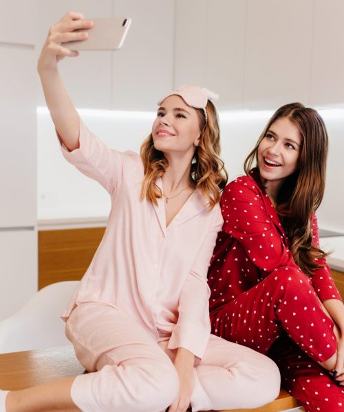 Romantic girls sitting on wooden table together and taking picture of themselves. Indoor shot of lovely ladies in pajamas making selfie in kitchen.